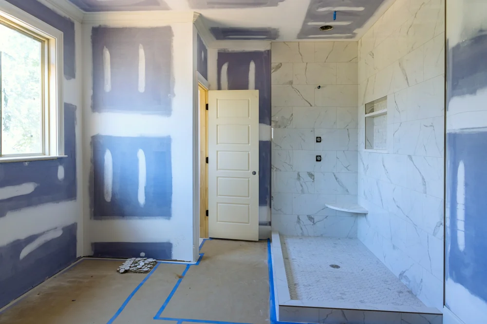 A bathroom under construction with unfinished drywall, a window, a white door, and a tiled shower area with a built-in bench. Blue tape marks are on the floor, and construction materials are present.
