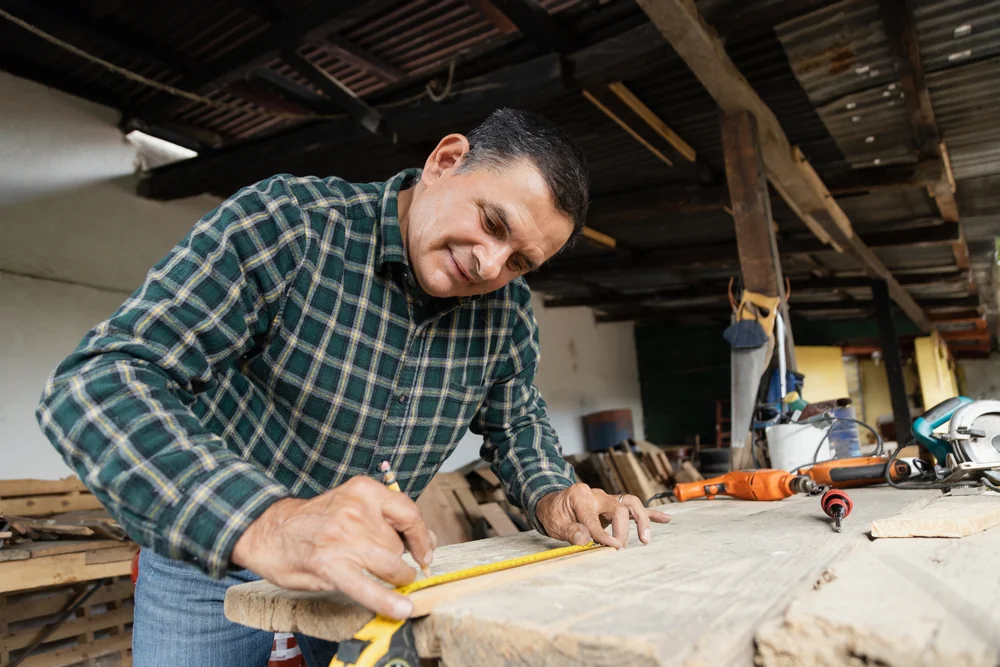 A man in a plaid shirt smiles as he measures and marks a wooden plank with a tape measure inside a workshop with various tools on the table.