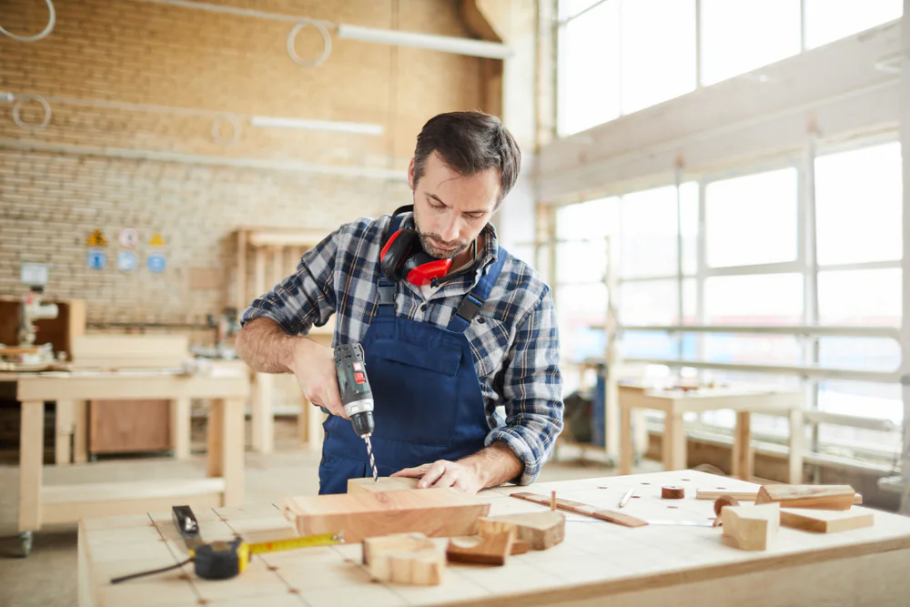 A man in a plaid shirt uses a power drill on a piece of wood at a workbench in a bright, spacious woodworking shop. Various tools and pieces of wood are scattered on the table.