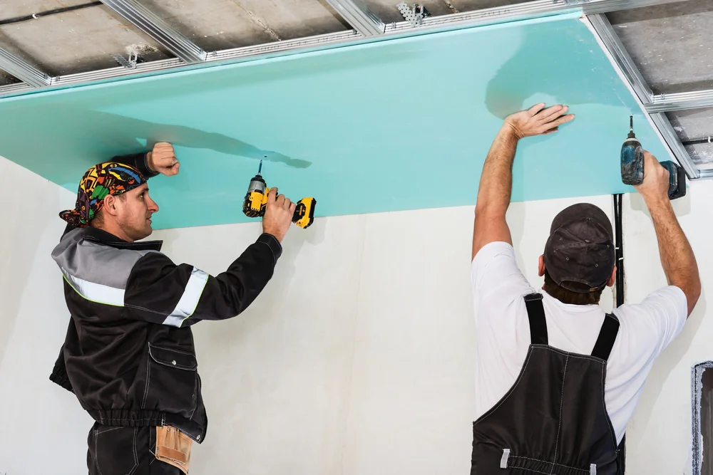 Two workers in overalls use electric drills to install a large sheet of drywall or plasterboard onto a ceiling, standing on the floor and securing it to a metal frame structure.