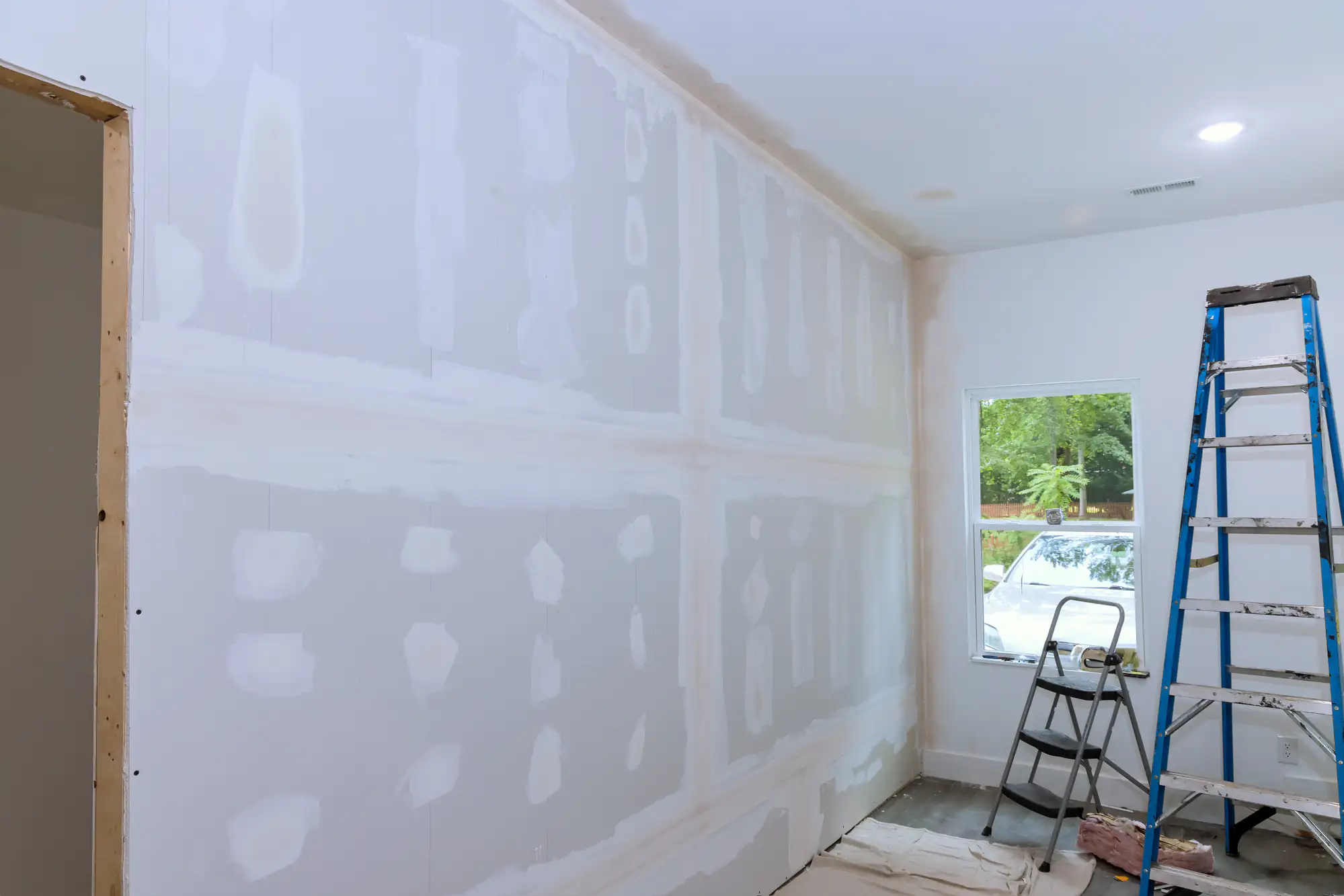 Room under construction with unfinished drywall, visible joint compound on seams, a window letting in daylight, and a ladder and step stool placed near the wall.