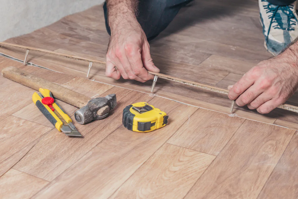A person measures and marks a wooden floor with a ruler and nails; a hammer, yellow tape measure, and utility knife are placed nearby on the floor.