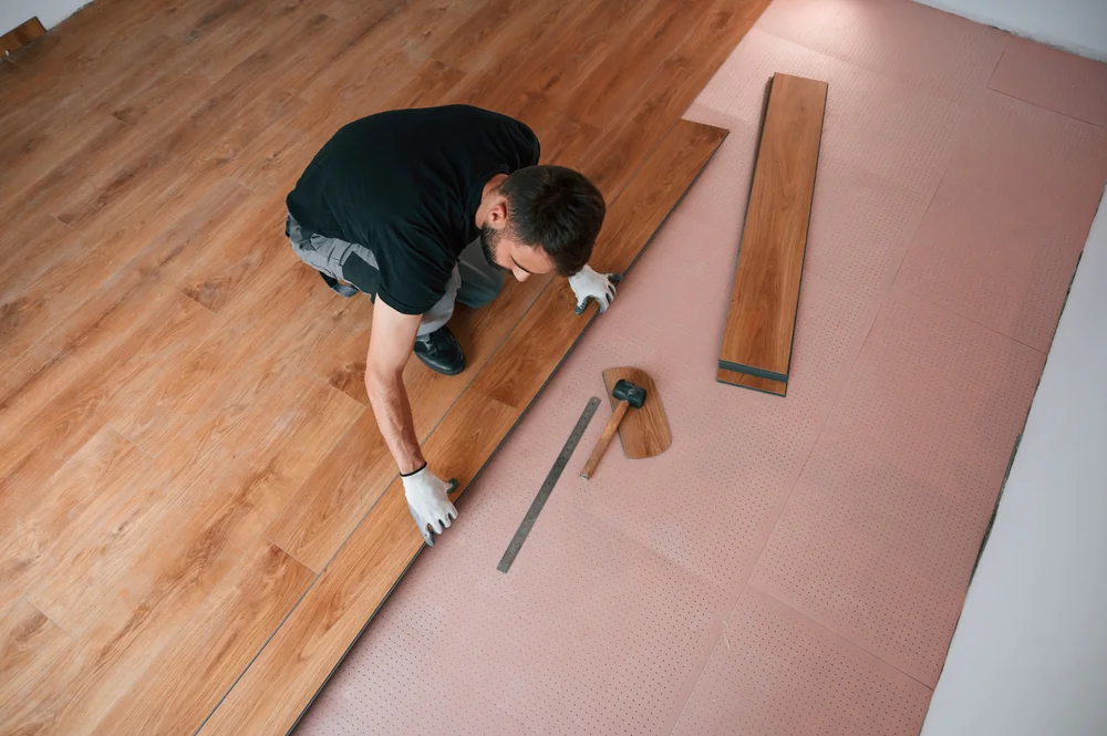 A man wearing gloves installs laminate wood flooring over a pink underlayment, using tools like a ruler, mallet, and tapping block, as he carefully fits a floorboard into place.