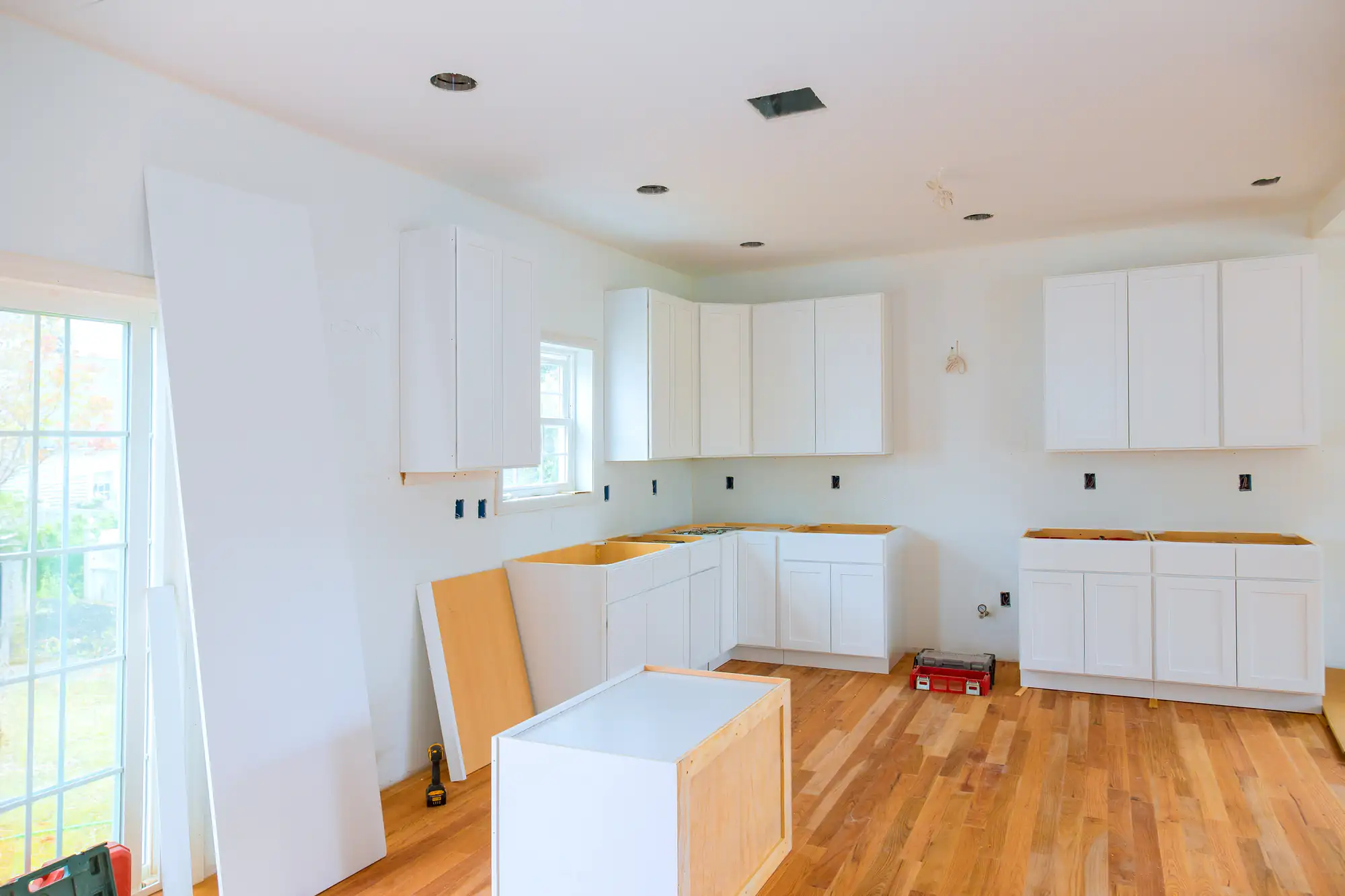 A kitchen under renovation with white cabinets being installed, an unfinished island in the center, tools and materials on the wooden floor, and natural light coming in from a sliding glass door.
