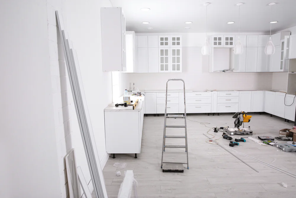 A modern kitchen under renovation with white cabinets, a ladder in the center, and various tools and construction materials scattered on the light wood floor. The walls and cabinetry are white.