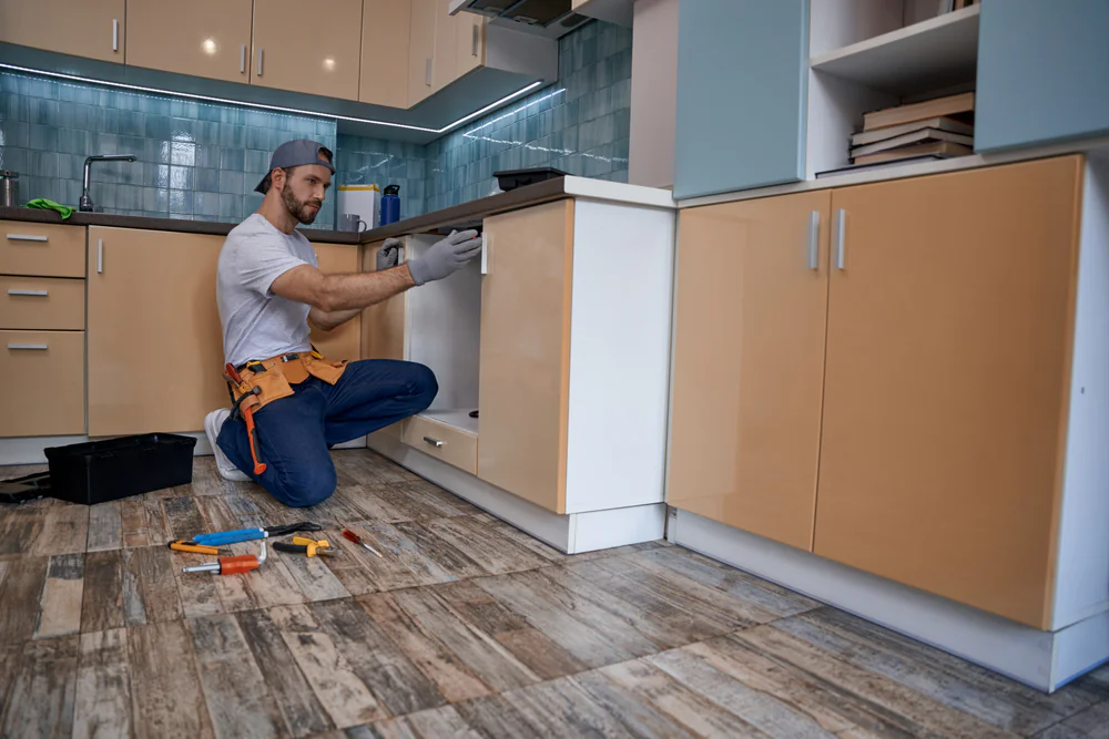 A man wearing a cap and tool belt kneels on a kitchen floor, using tools to repair a cabinet. Various tools and a toolbox are scattered nearby in a modern kitchen with tan lower cabinets and blue backsplash.