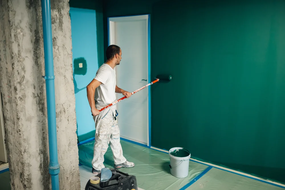 A man in white work clothes uses a paint roller to paint a wall dark green. A bucket of paint is on the plastic-covered floor nearby, and there is an unfinished concrete column in the room.