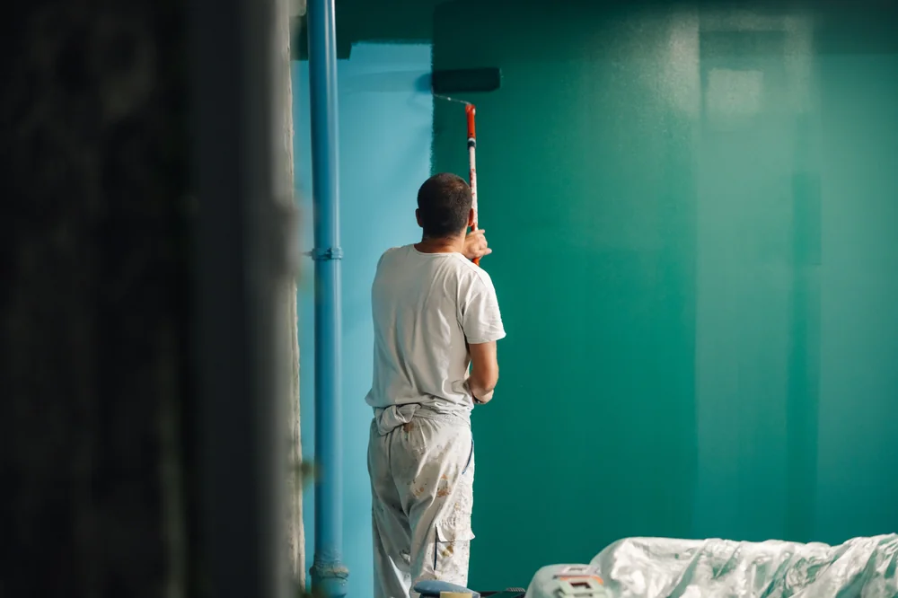 A person wearing white clothes is painting a wall teal using a roller brush. The floor is partially covered with a plastic sheet, and the room appears to be in the process of being renovated.
