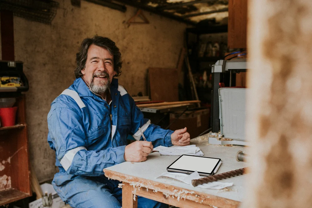 A smiling man in a blue work jumpsuit sits at a cluttered workbench in a workshop, holding papers. An open tablet and various tools are on the table.