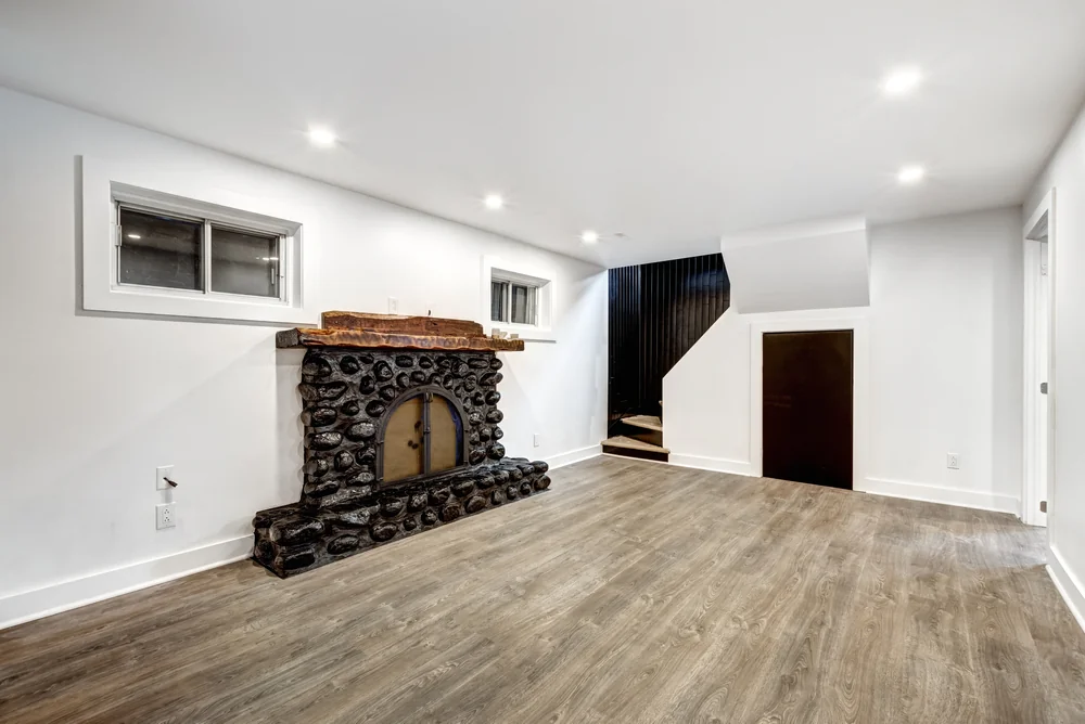 A spacious room with wood-style flooring, white walls, a dark stone fireplace, two small windows, recessed ceiling lights, and a staircase with a black railing next to a small door under the stairs.