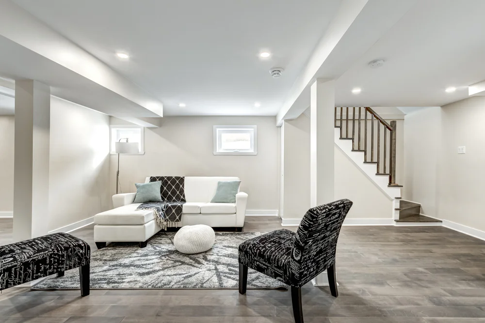 Modern finished basement with light walls, dark wood floors, a white sofa, patterned black chairs, a white ottoman, a gray rug, a pouf, and a staircase with a dark banister in the background.