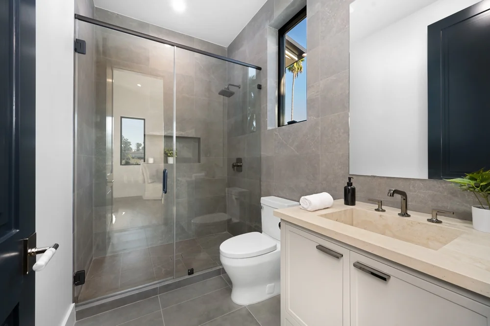 Modern bathroom with gray tile, glass-enclosed shower, white toilet, and beige countertop vanity with sink, faucet, soap dispenser, small plant, and large mirror; window provides natural light.