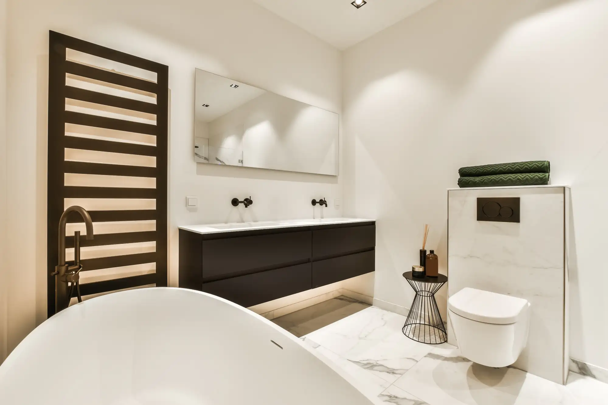 Modern bathroom by a general contractor Suffolk County, NY, featuring a freestanding bathtub, double sink vanity, large mirror, wall-mounted toilet, black towel warmer, and green towels on a marble shelf. The decor is minimalist and elegant.