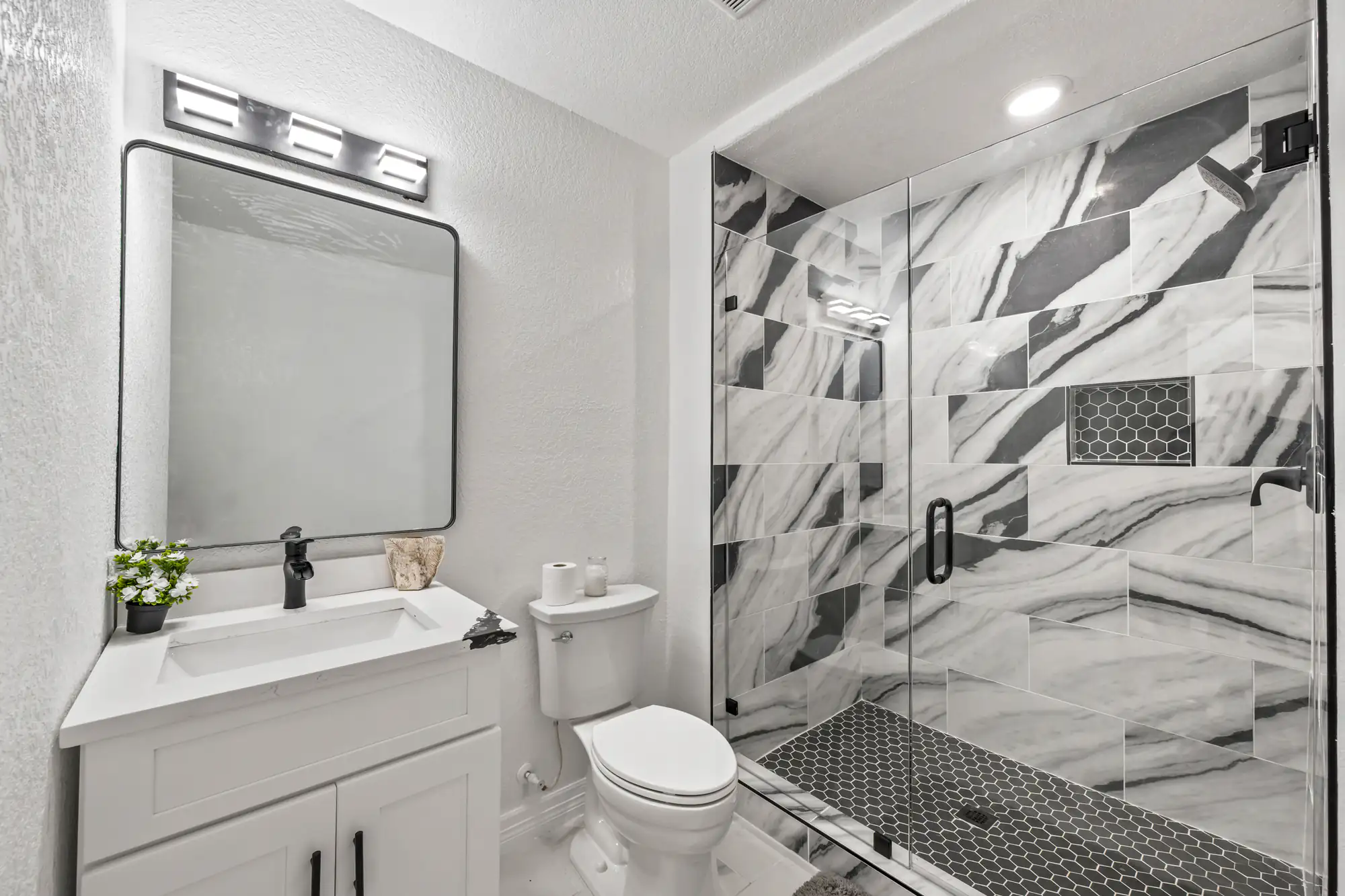 Modern bathroom by a general contractor Suffolk County, NY, featuring a white vanity, rectangular mirror, toilet, and glass-enclosed shower with striking black and white patterned tiles and hexagonal flooring. A small plant and towel add charm.