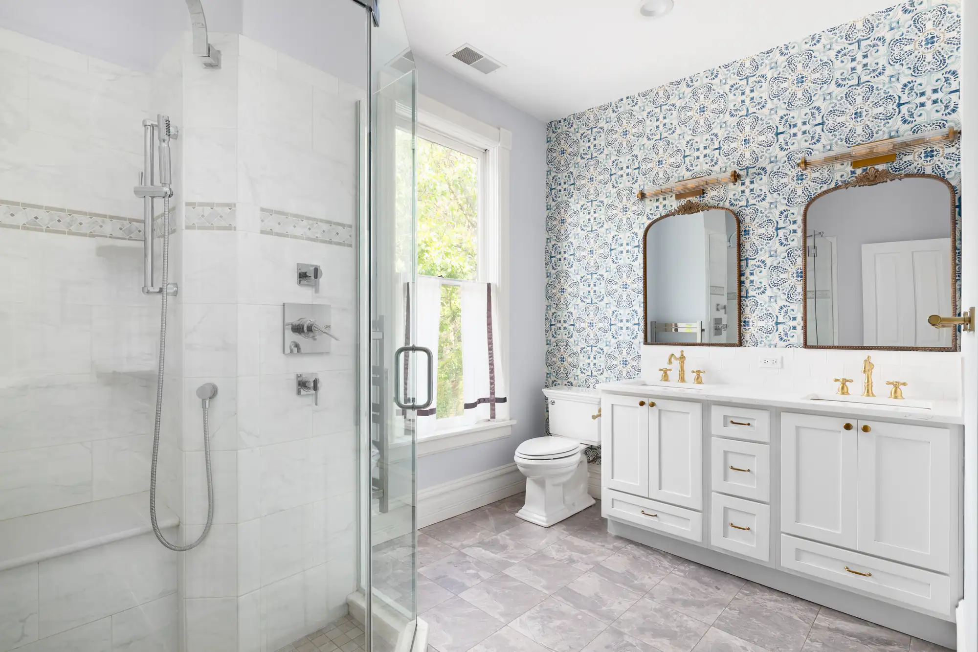 Bright bathroom with a glass shower, marble walls, double vanity and gold fixtures. A general contractor in Suffolk County, NY can bring your vision to life with a blue-and-white accent wall, large window, and gray tile floor.