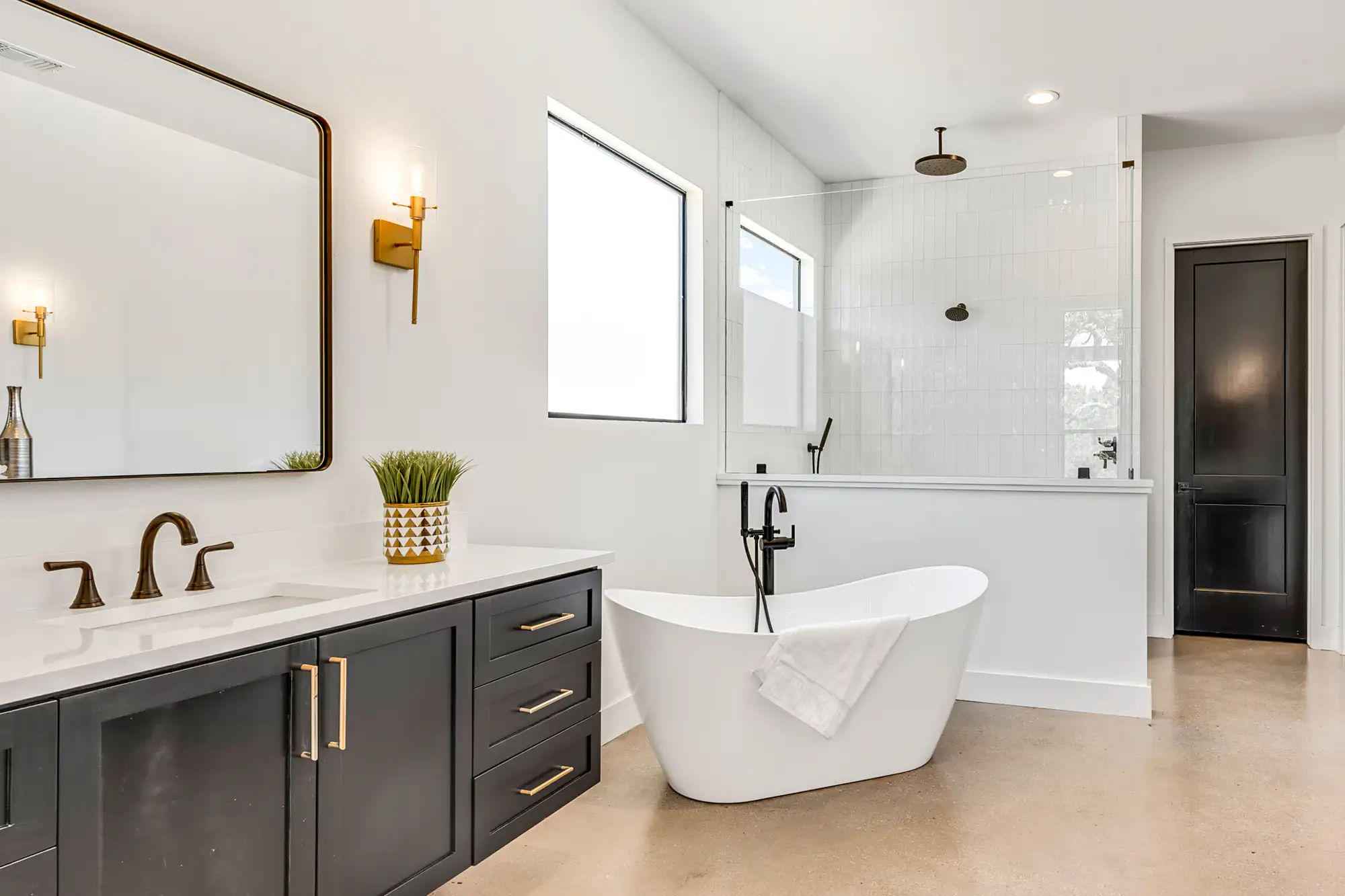Modern bathroom by a general contractor Suffolk County, NY, with a freestanding white tub, black fixtures, double sink with dark cabinets, large mirror, wall sconce lighting, glass-enclosed shower, and minimal decor on a light floor.