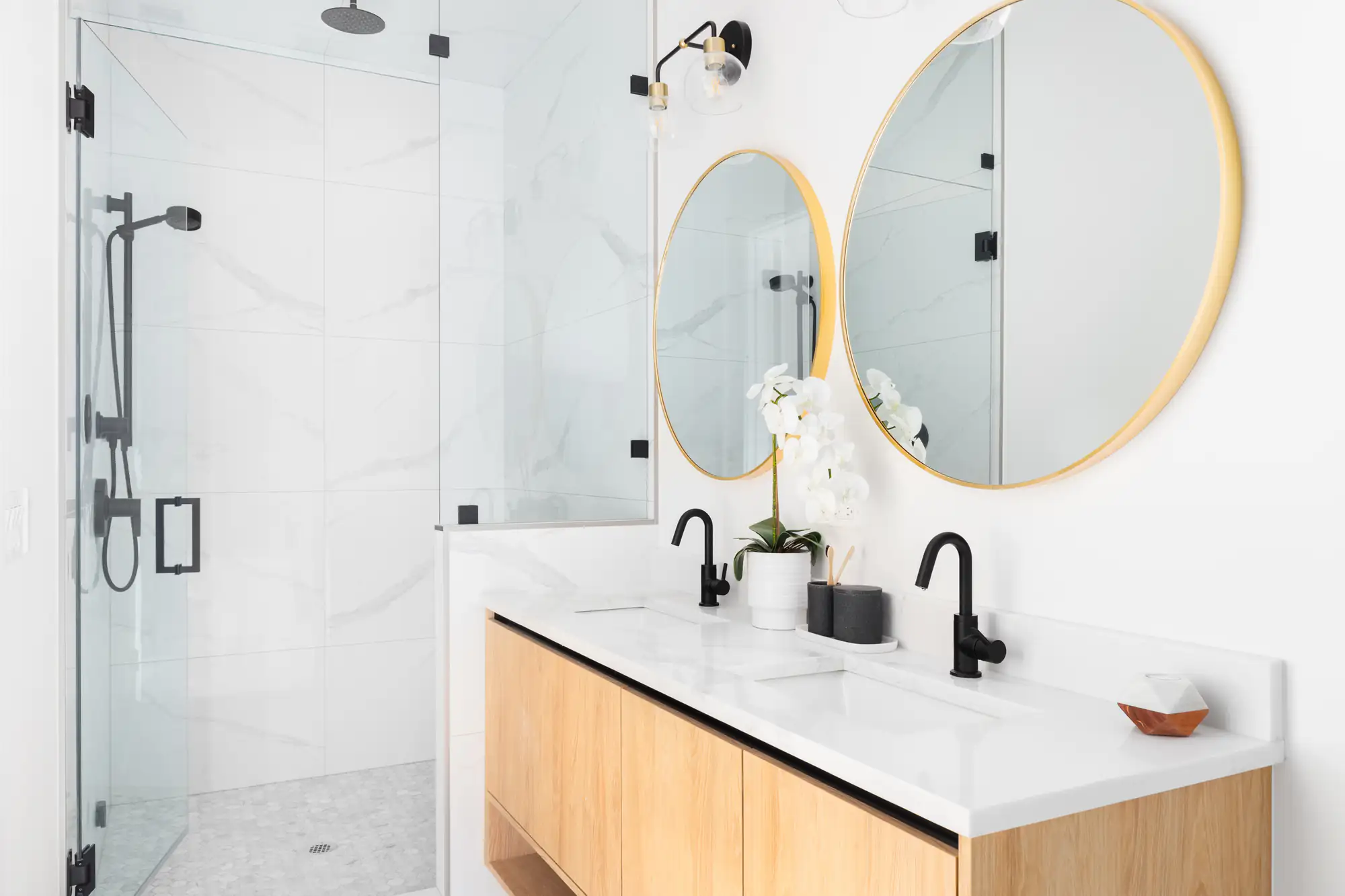 Modern bathroom with a glass shower, black fixtures, double sink vanity with light wood cabinets, two round gold-framed mirrors, white countertop, and a white orchid plant&mdash;expertly crafted by a general contractor in Suffolk County, NY.