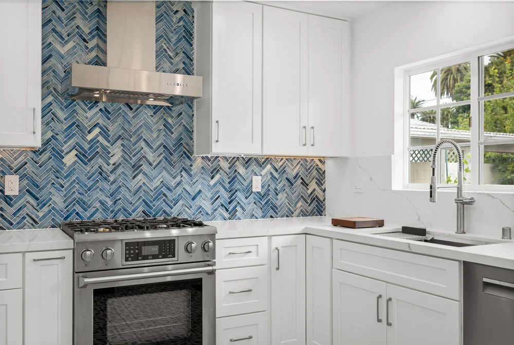 Modern kitchen with white cabinets, stainless steel stove and hood, blue herringbone tile backsplash, marble countertops, and a large window above a sink with a pull-down faucet.