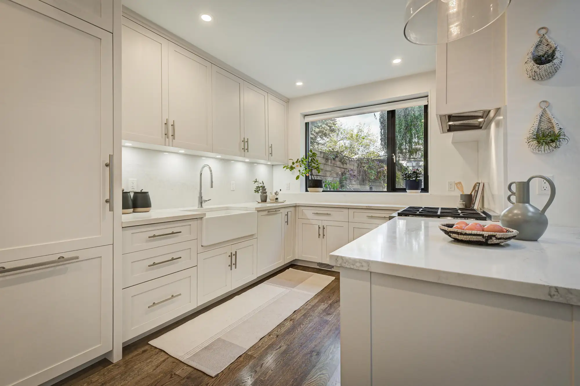 Modern kitchen with white cabinets, farmhouse sink, gas stove, under-cabinet lighting, large window with a view of greenery, decorative plants, and wooden flooring. Neutral decor and natural light create a bright, inviting space.