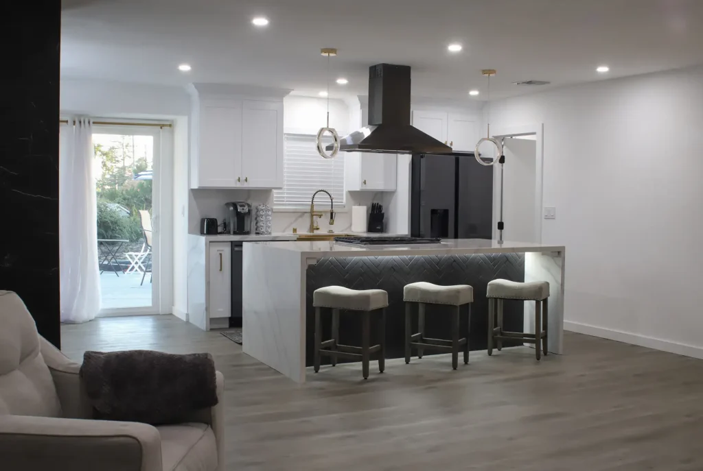 Modern kitchen with a central island, three bar stools, white cabinets, black appliances, and a sleek range hood—crafted by top General Contracting Suffolk County, NY experts. Sliding glass doors open to a patio with outdoor seating.
