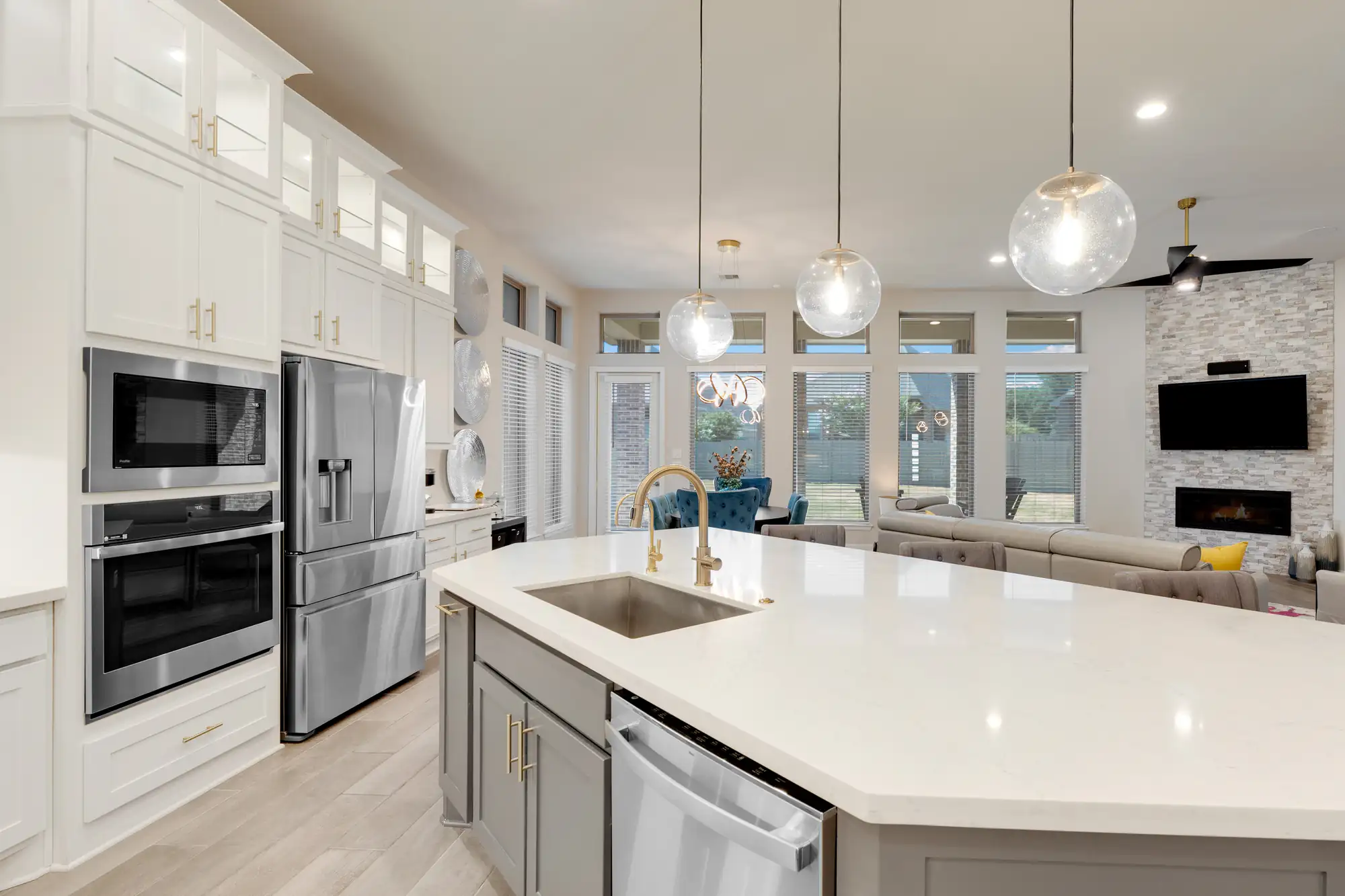 Modern kitchen with a large island, built-in sink, gray cabinets, stainless steel appliances, pendant lights, and an open view into a bright living area with windows, a couch, and a wall-mounted TV.
