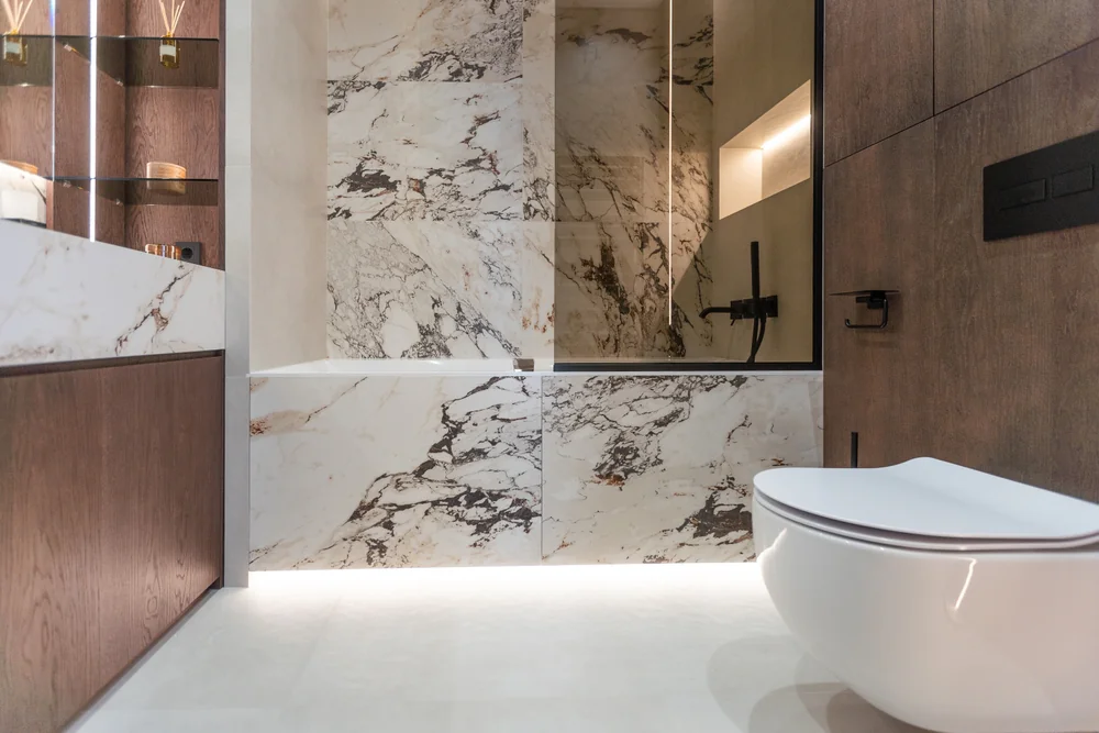 Modern bathroom with a floating white toilet, marble wall and bathtub, wood-paneled cabinets, glass shower partition, and soft under-cabinet lighting creating a clean, luxurious look.