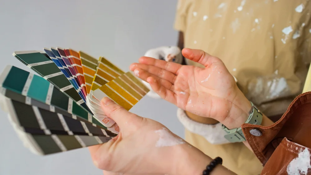 Two people with paint-stained hands hold and look at a fan of paint color swatches, considering various shades of green, yellow, orange, and red against a neutral background.