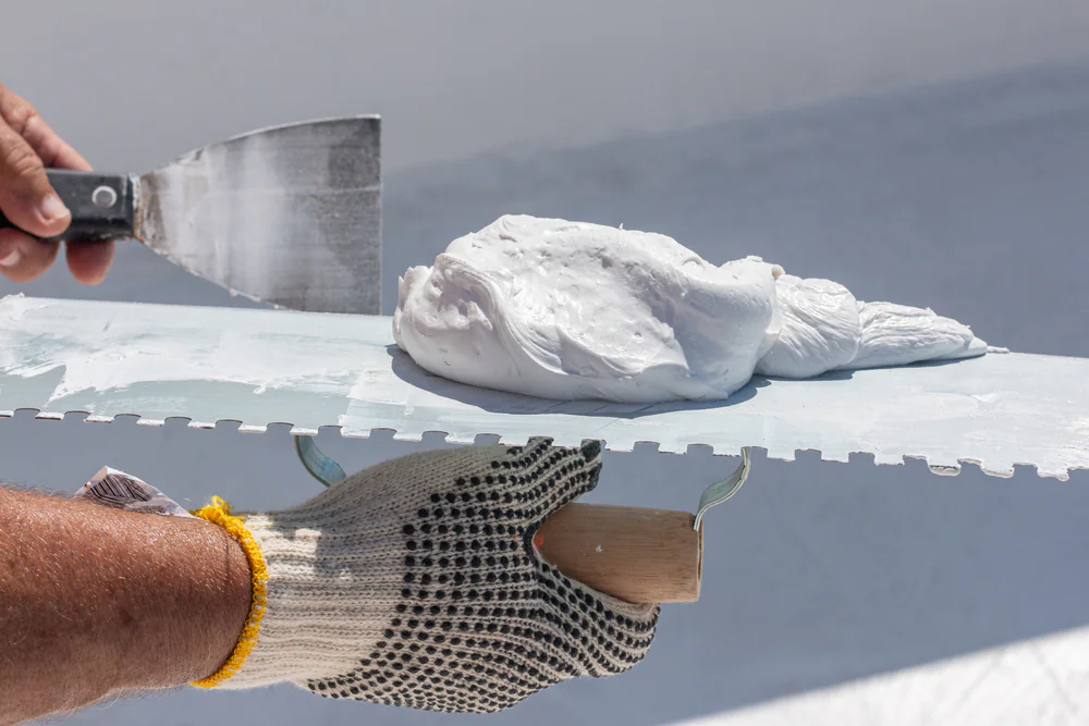 A person wearing a protective glove holds a notched trowel with a mound of white plaster or adhesive, while another hand holds a putty knife with some of the material on it.