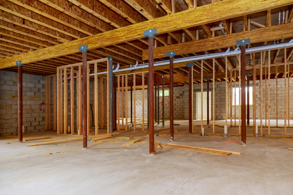 Interior of a building under construction showing exposed wooden framing, support beams, concrete floor, and unfinished cinder block walls. The ceiling joists, wiring, and plumbing are also visible.