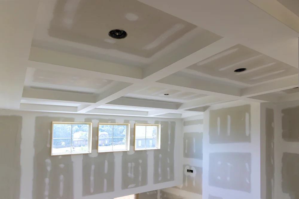 Unfinished drywall ceiling and walls with recessed lighting, featuring a coffered design. Multiple rectangular sections are visible, along with several windows letting in natural light.