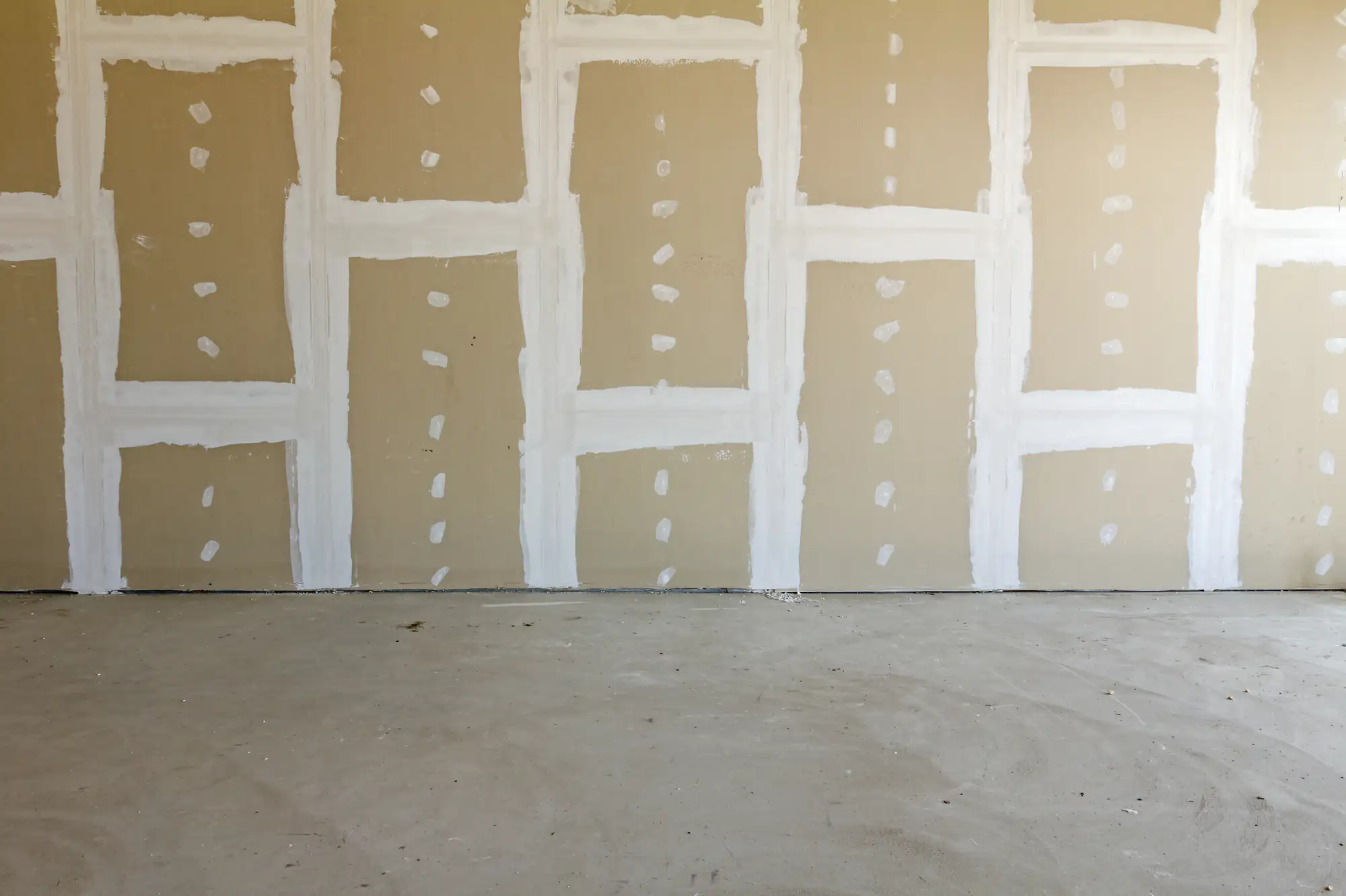 A bare drywall wall with visible seams and joints covered in white joint compound, set above an unfinished concrete floor in a construction or renovation setting.