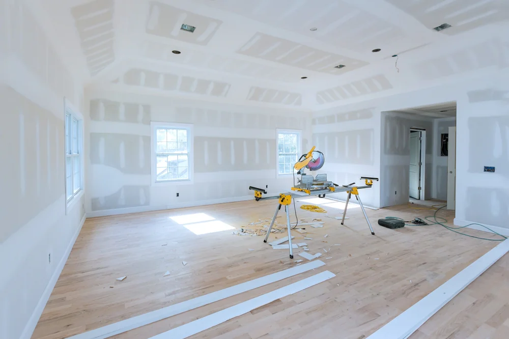 A bright, unfinished room with white drywall, wooden floors, and a yellow miter saw on a stand surrounded by wood planks and debris. Sunlight streams through two windows.