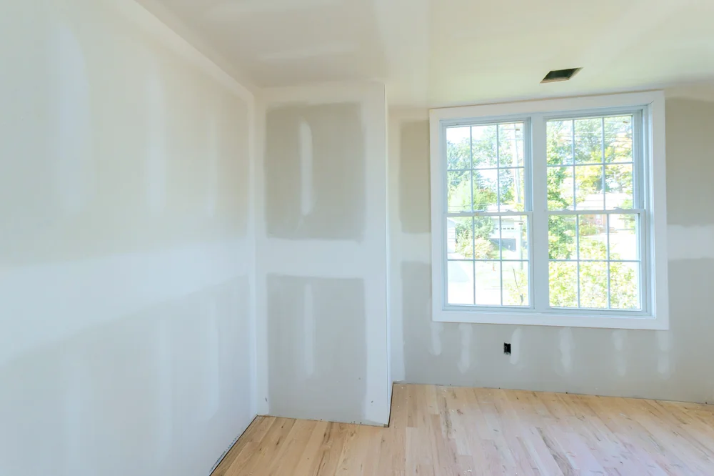 A bright, empty room with unfinished drywall, light wood flooring, and a large window letting in natural light. Trees and sky are visible outside the window.