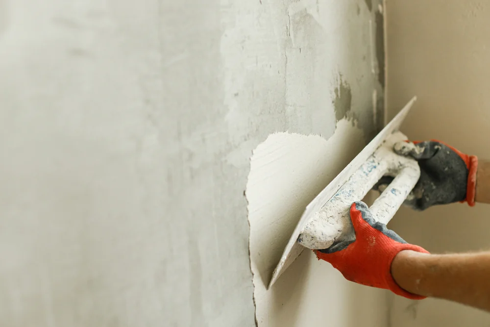 A person wearing orange gloves smooths plaster on a wall using a trowel, applying a fresh coat of plaster to the surface.