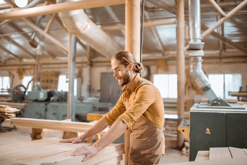A man wearing an apron works with wooden planks in a spacious, well-lit woodworking shop, surrounded by industrial machines and equipment.
