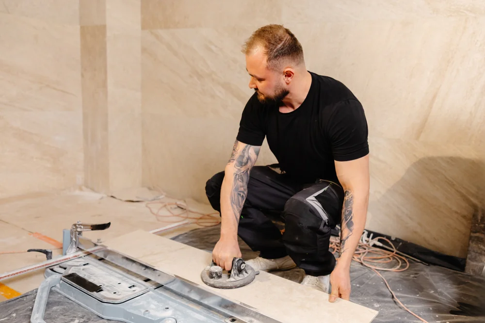 A man with tattoos wearing a black t-shirt and pants uses a tile suction cup lifter to handle a large ceramic tile in an indoor construction or renovation setting.