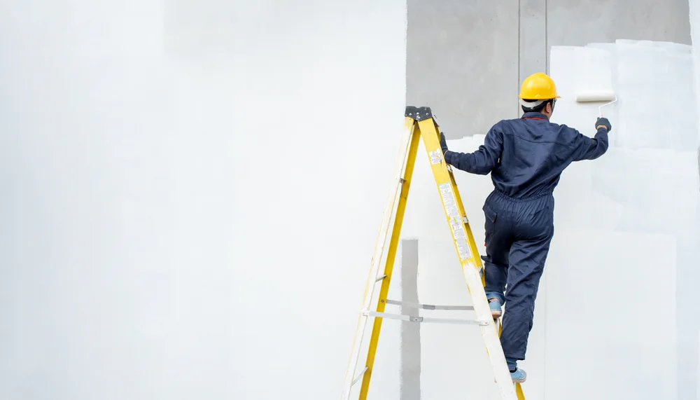 A person wearing a yellow hard hat and blue coveralls stands on a yellow ladder, painting a white wall with a paint roller. The background is mostly white with some unfinished gray areas.