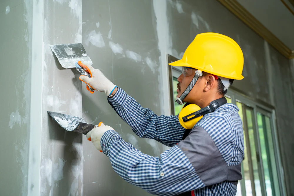 A construction worker wearing a yellow hard hat, safety goggles, and gloves is using two putty knives to apply plaster or spackle to a drywall surface inside a building.