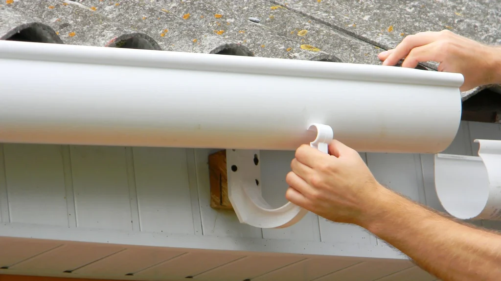 Hands install a white gutter system on a gray shingle roof edge, highlighting seamless gutter installation.