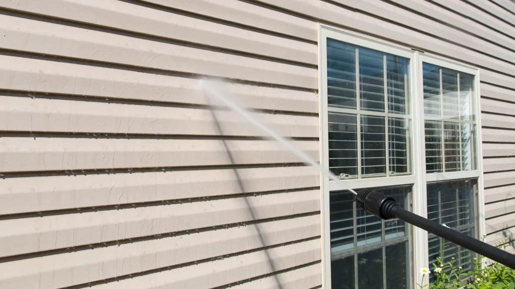 A power washer cleaning the beige house siding next to a window with a strong spray of water.