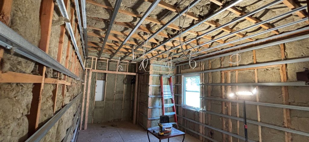 A room under renovation featuring metal wall studs, insulation, and electrical wiring from the ceiling.
